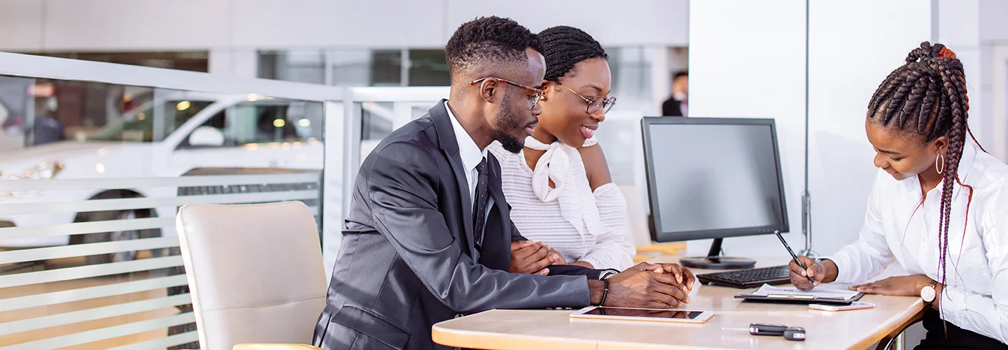 A group of three people at a table in a car dealership, reviewing paperwork and discussing car purchases.