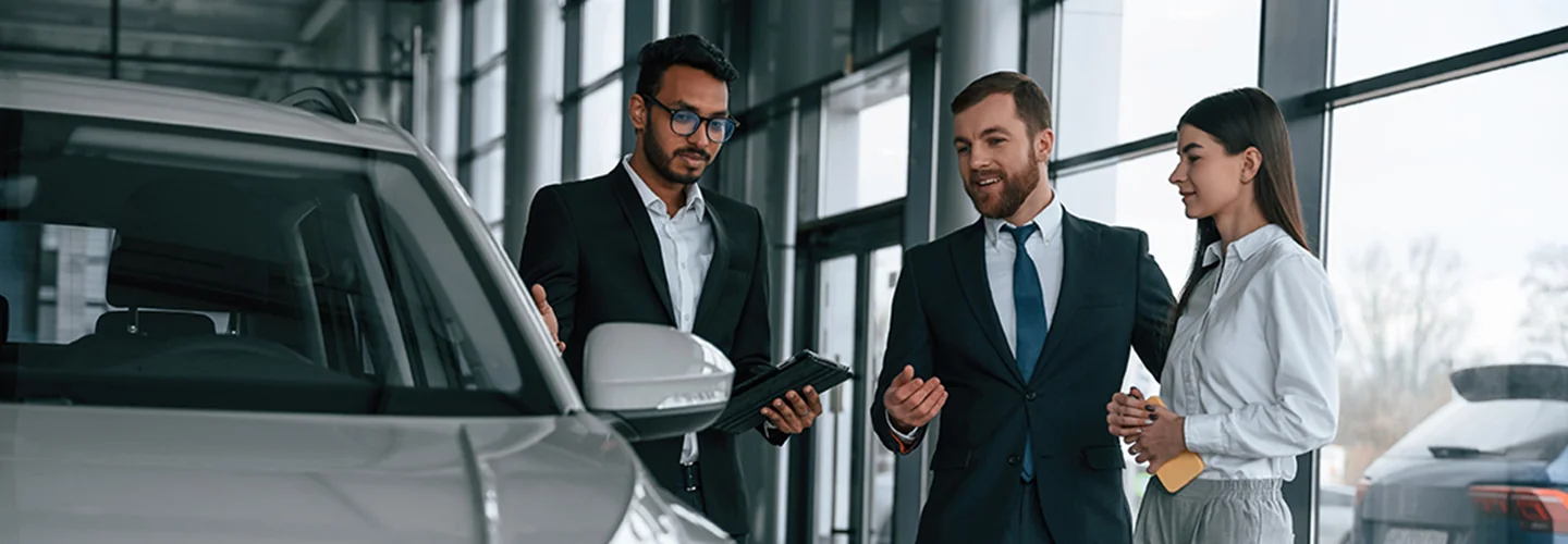 Three business professionals standing together in front of a parked car, dressed in formal attire.