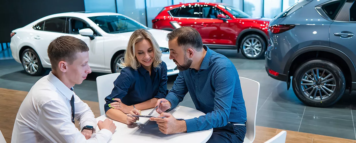 Dealership sales consultant assisting customers with vehicle financing and purchase paperwork in a modern automotive showroom