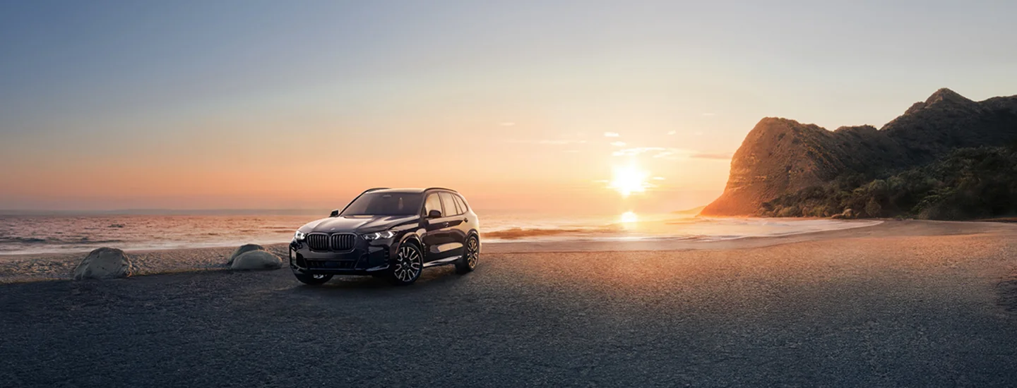 A parked black 2026 BMW X5 at the ocean with the sun setting