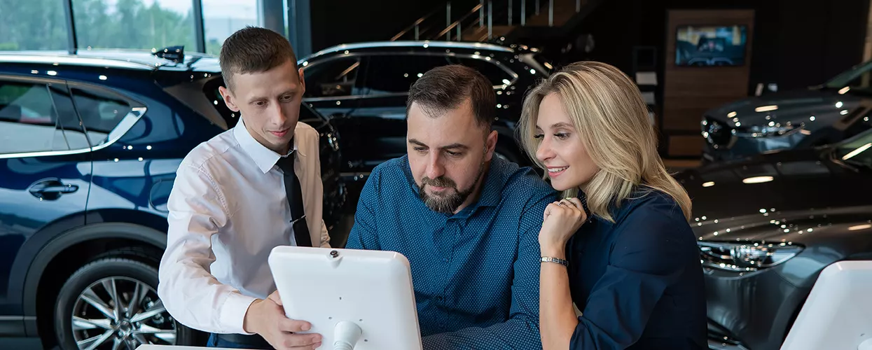 Car dealership sales consultant helping customers review vehicle features and pricing on a digital display in a modern showroom