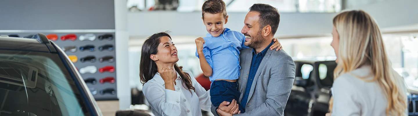man holding a child with another person cherring the purchase of their new car with the salesperson looking on