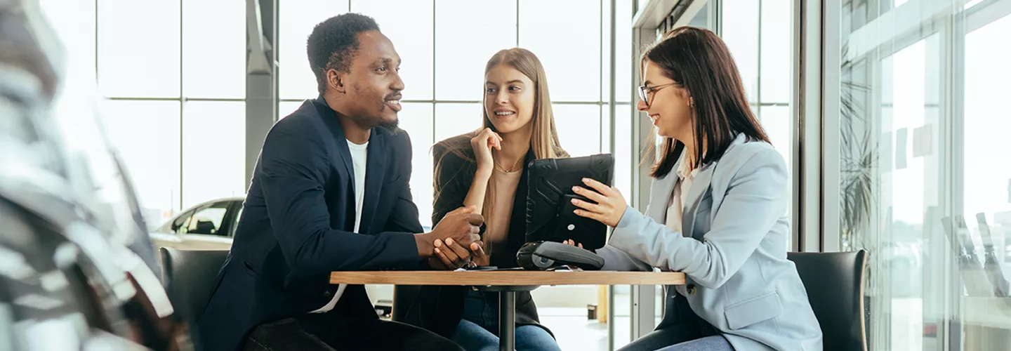 Three people sitting at a table in a car dealership talking about a contract