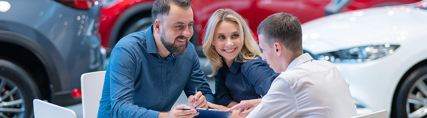 couple smiling talking with car dealer salesperson sitting at desk in showroom