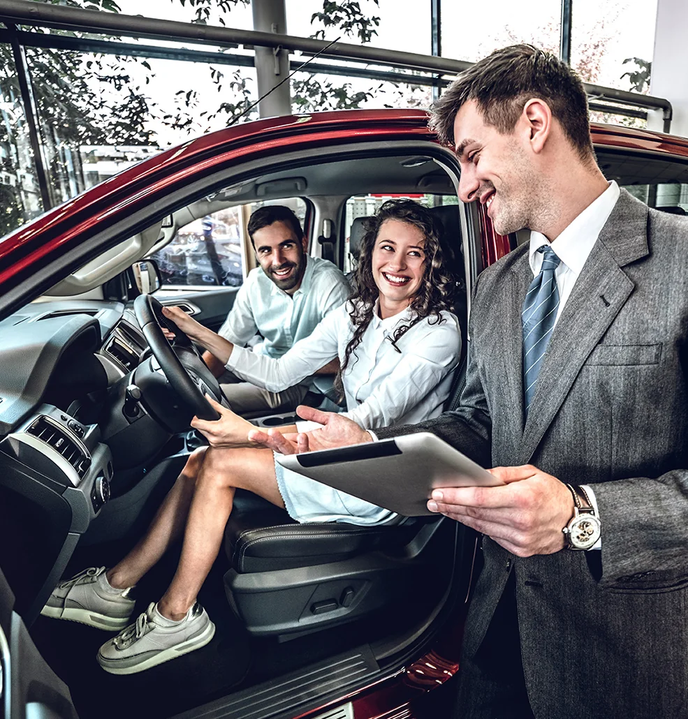A man and woman seated in a car, the woman is holding a map while the man drives.