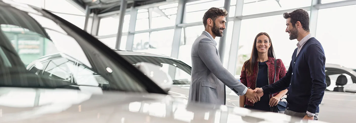 Two people shaking hands with another looing on in car dealership