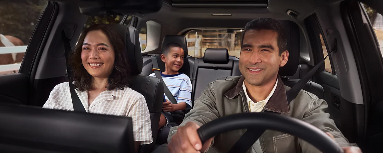 A family of four is driving in a car, featuring a man and woman in the front seats with children in the back.