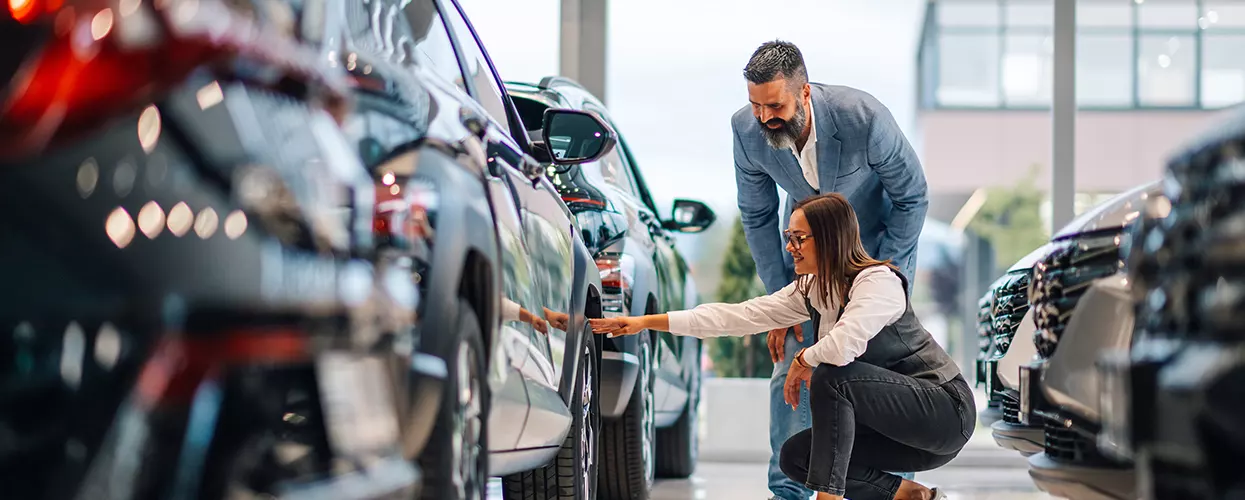 one person kneeling by tire of car at a dealership with another standing by going over details of new car