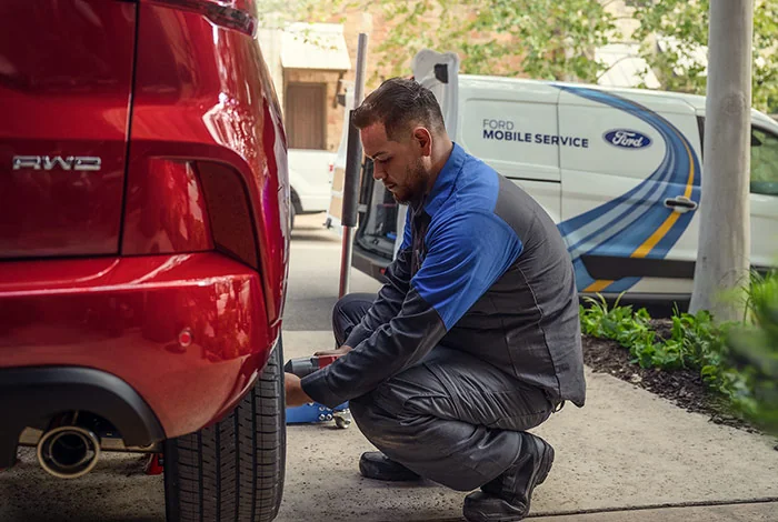 Ford Mobile Service Mechanic working on an AWD red Ford. He has his tire wrench with a jack and his Ford Mobile Service vehicle parked at the curbside.