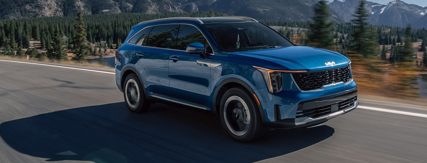 Front side view of a blue 2025 Kia Sorento Hybrid driving on a road next to a forest