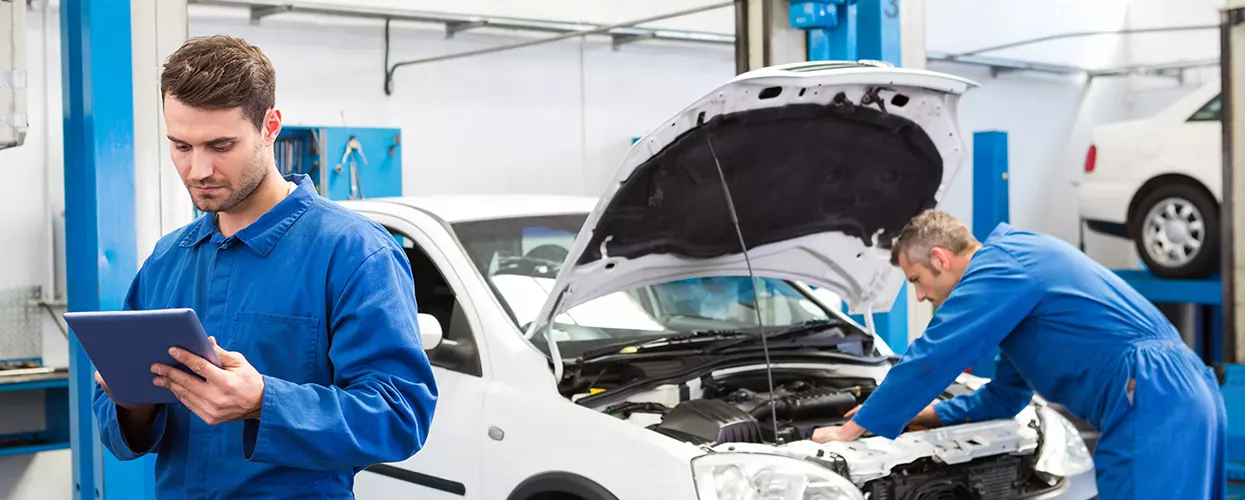 Service mechanic looking at a tablet and a vehicle with the hood up behind him