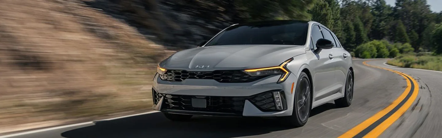 Front view of a gray 2025 Kia K5 Specs driving on a road by a forest