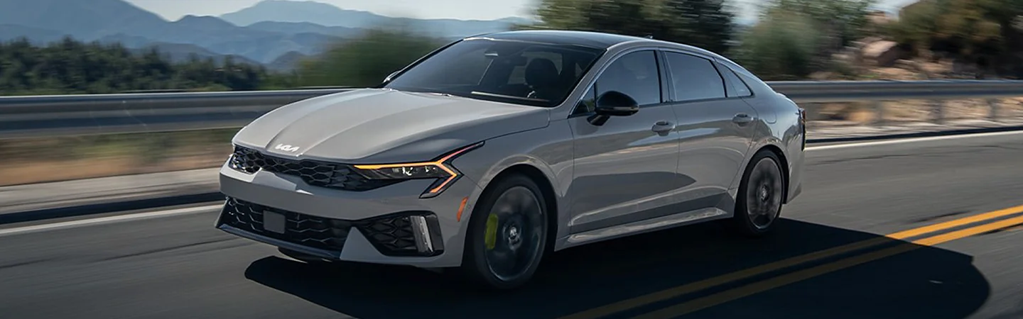 Side view of a gray 2025 Kia K5 Driving on a road with mountains in the background