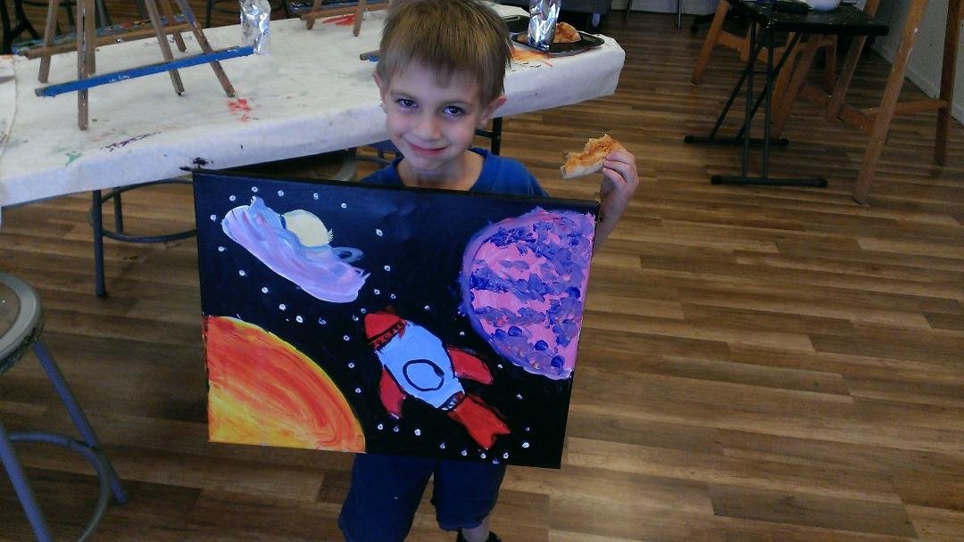 My 5 year old grandson, Jadon, showing off his painting of a rocket ship off to some planet (maybe not even in our solar system), and holding a slice of pizza in his hand. This was taken at an art studio at a birthday party.