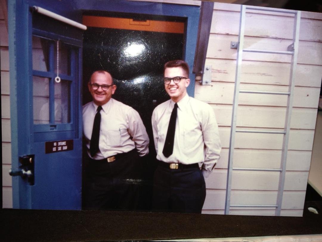 I ran across this photo while organizing my desk. Who is the guy on the right, what is he doing and why is he smiling? And where is the photo taken? No prize, just wondering if you might know.