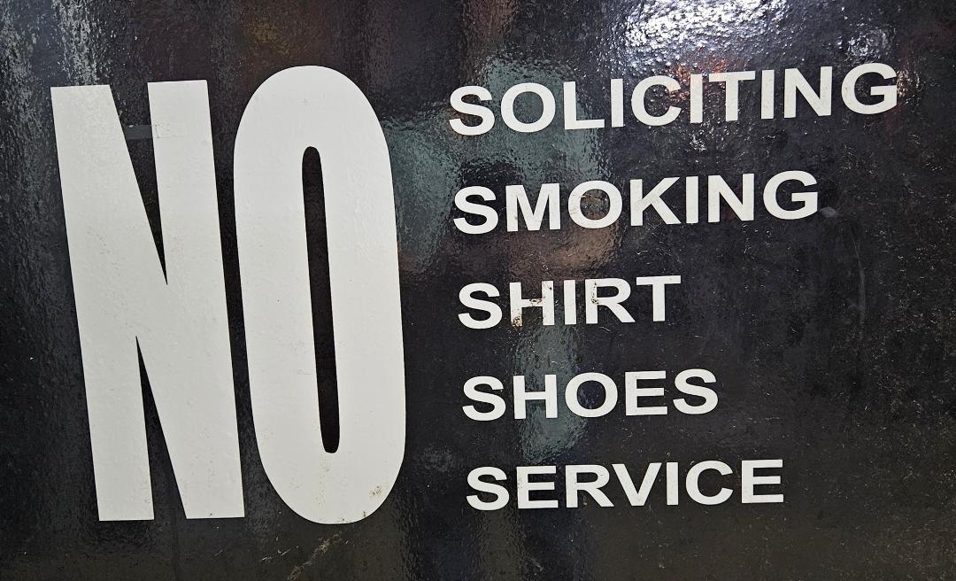 Grammatically, this sign either means patrons may not wear shirts or shoes and must not provide service, or they must smoke and solicit.