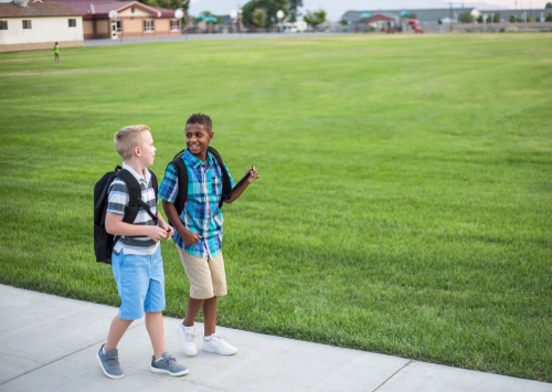 Two diverse school kids walking home together after school and talking together. Back to school photo of diverse school children wearing backpacks.
