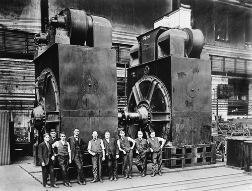 Men in suits in front of two massive electric generators in a factory, around 1920