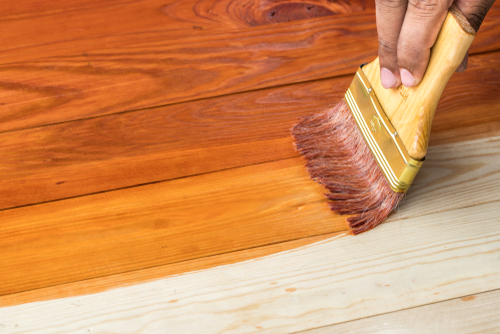 A hand holding a brush applying a coat of varnish on a wooden surface.