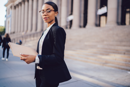 Portrait of confident African American lawyer in stylish eyeglasses and formal apparel holding folder documents in hands while looking at camera. 