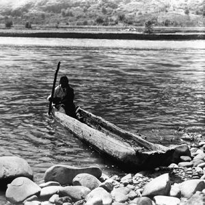 man with pole maneuvering dugout canoe to rocky shore.
