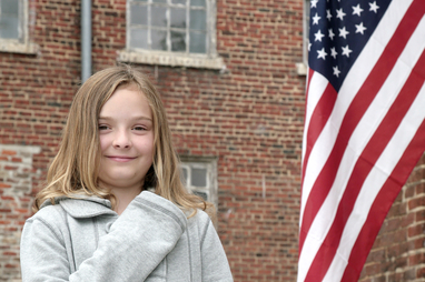 one young cute child with hand on her heart near an American Flag outdoors