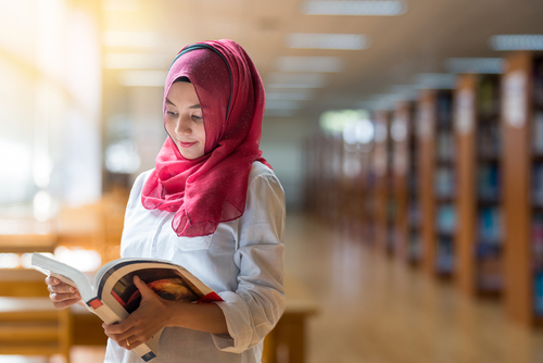 Muslim girl reading book with hijab on blur library background.