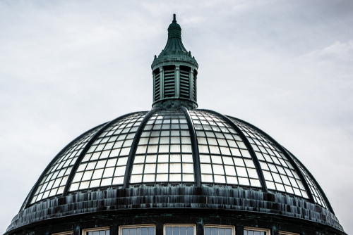 Isolated aviary dome with square glass paneling and round pointed cupola projecting from top center. Gray-blue cloudy sky fills background. Cascading waterfall runs along bottom circumference.
