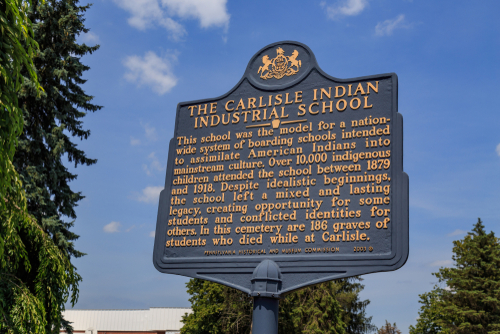 Photograph shows Eskimos (Inuit) constructing an igloo with blocks of snow as children and dogs stand by.