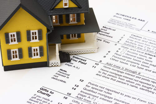 Cropped image of businessman calculating tax by model house and stacks of coins on table.