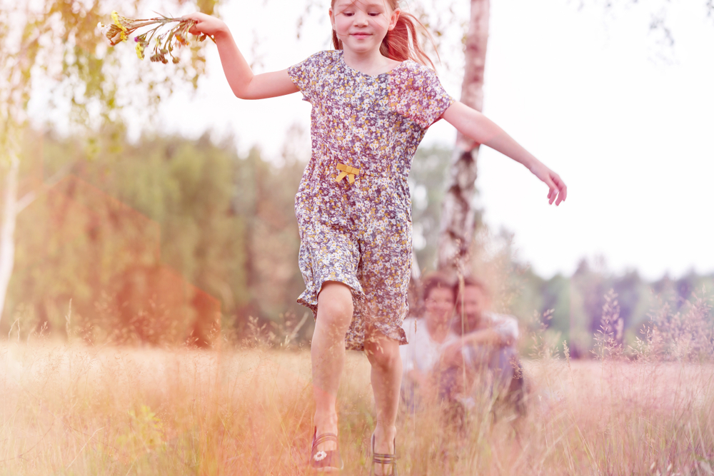 Happy young girl running and playing while her parents are sitting on a field behind her.