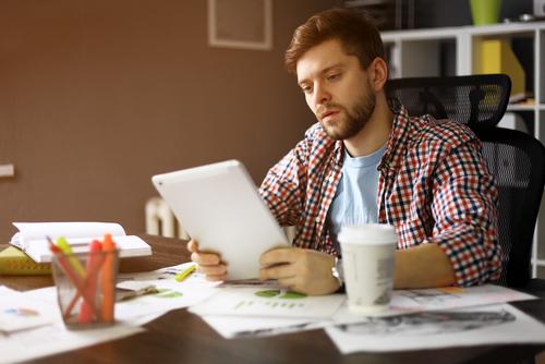 Thoughtful male person looking to the digital tablet screen while sitting in modern loft interior at the table, experienced entrepreneur reading some text or electronic book at the office.