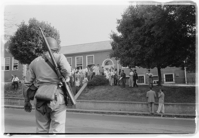 Photograph shows an armed member of the National Guard observing Clinton High School as students stand on its steps and lawn.