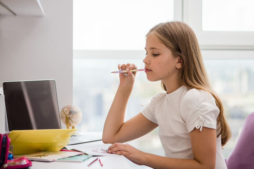Girl thinking while holding a pencil in her mouth and using a laptop computer.