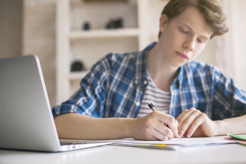 A casual young man writing essay at desk with a laptop close by.