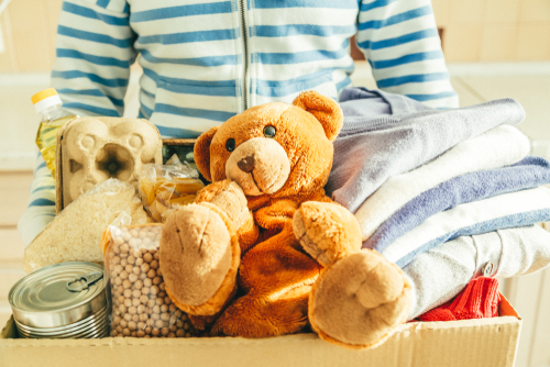 Donating concept - female holding food, clothes, toys in cardboard, kitchen background