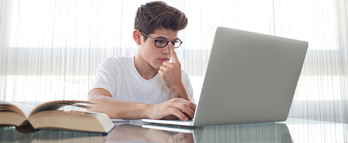 Panoramic view of teenager doing his homework at home, wearing reading glasses using a laptop computer, sitting at desk with window, home living room interior. Technology student lifestyle indoors.