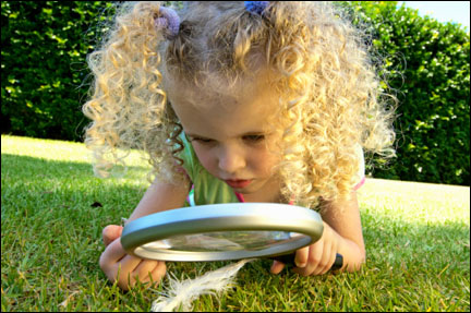 Girl (5) lying on grass, looking at feather through a magnifying glass.
