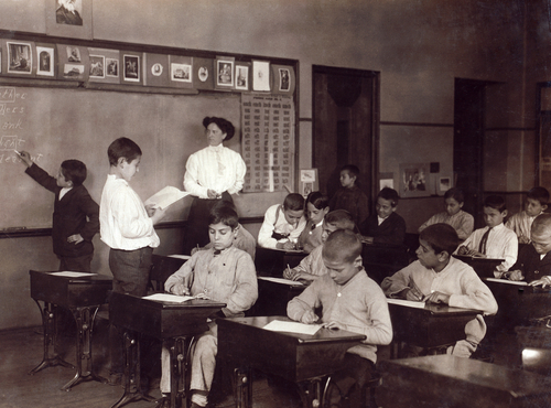 Photograph shows Eskimos (Inuit) constructing an igloo with blocks of snow as children and dogs stand by.