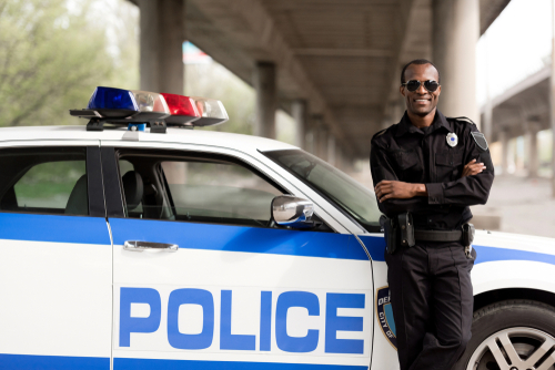 handsome african american police officer with crossed arms leaning back on car and looking at camera