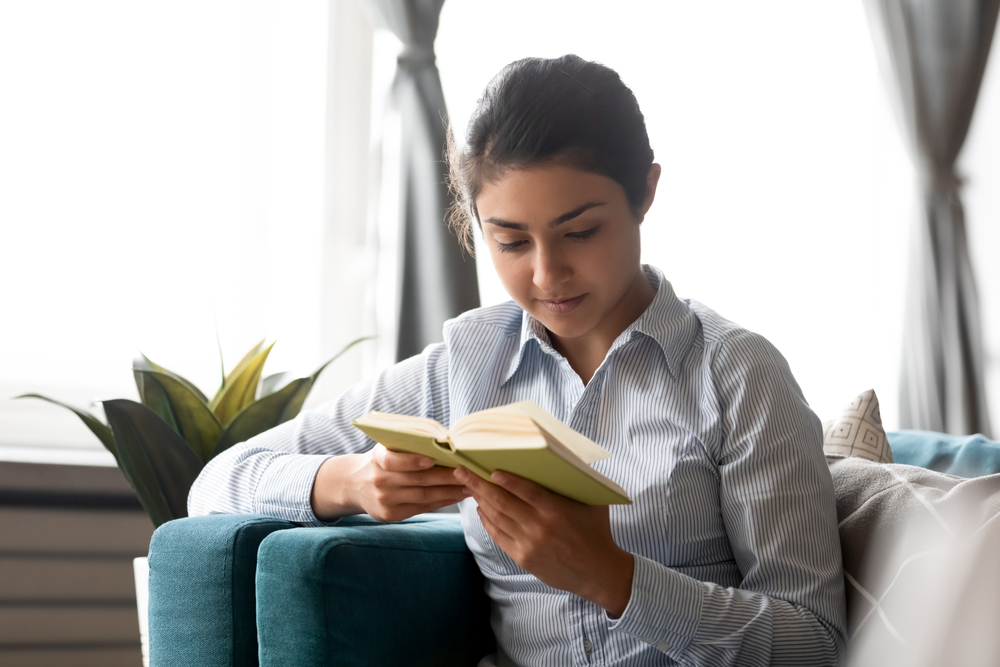 Focused young woman reading book while sitting on a cozy sofa.