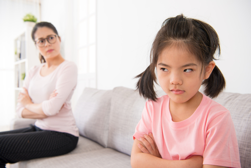 A young girl with her arms crossed looks away from her mother, whose arms are also crossed