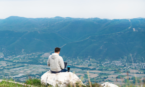 Young man with laptop sitting on top if mountain working online.