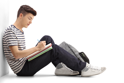 Teen student writing in a notebook and leaning against a wall isolated on white background.