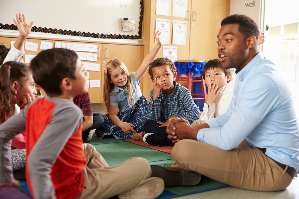 Elementary school kids and teacher sit cross legged on floor</strong>