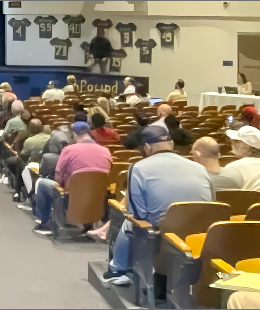 A large crowd is seated in an auditorium facing a panel of speakers on stage, with banners and jerseys decorating the walls.