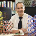Omar Yaghi seated at a desk wearing a striped shirt and dark tie, surrounded by colorful three-dimensional molecular framework models. Bookshelves and a computer monitor are visible in the background.