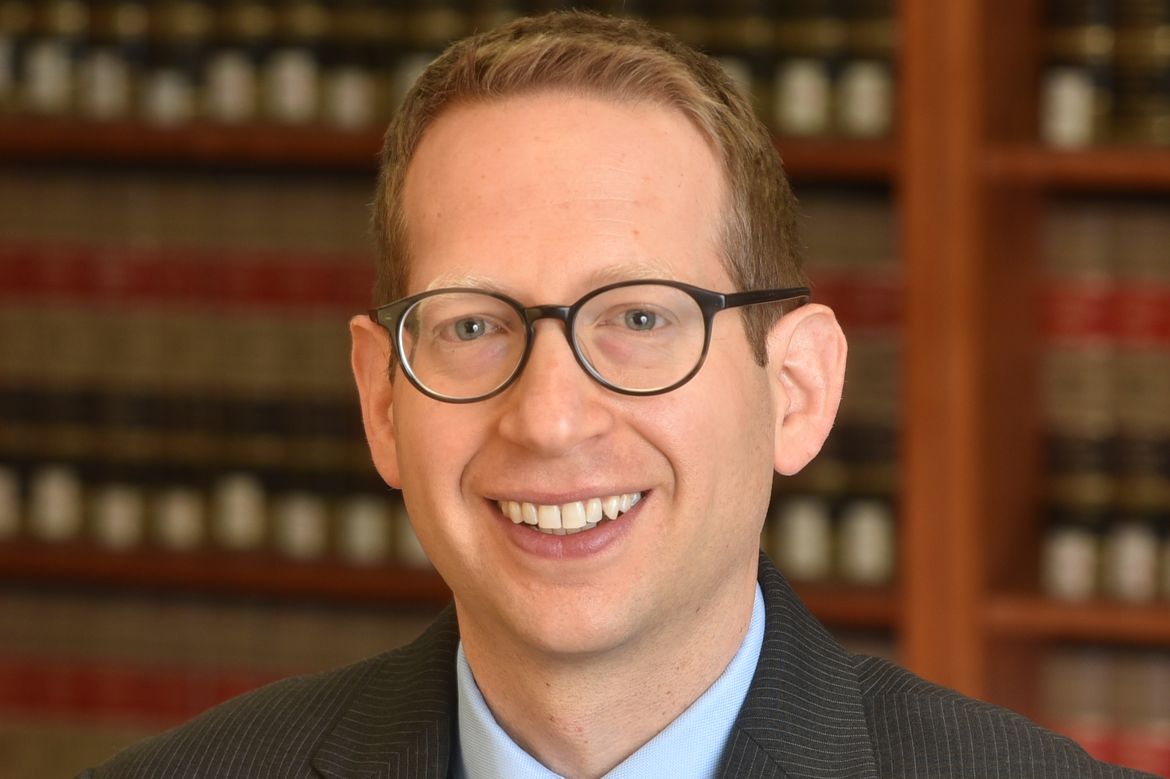 Jon Michaels in a dark gray suit and solid gray tie sits in front of a bookcase. The shelves are filled with matching black, gold and red-spined books.