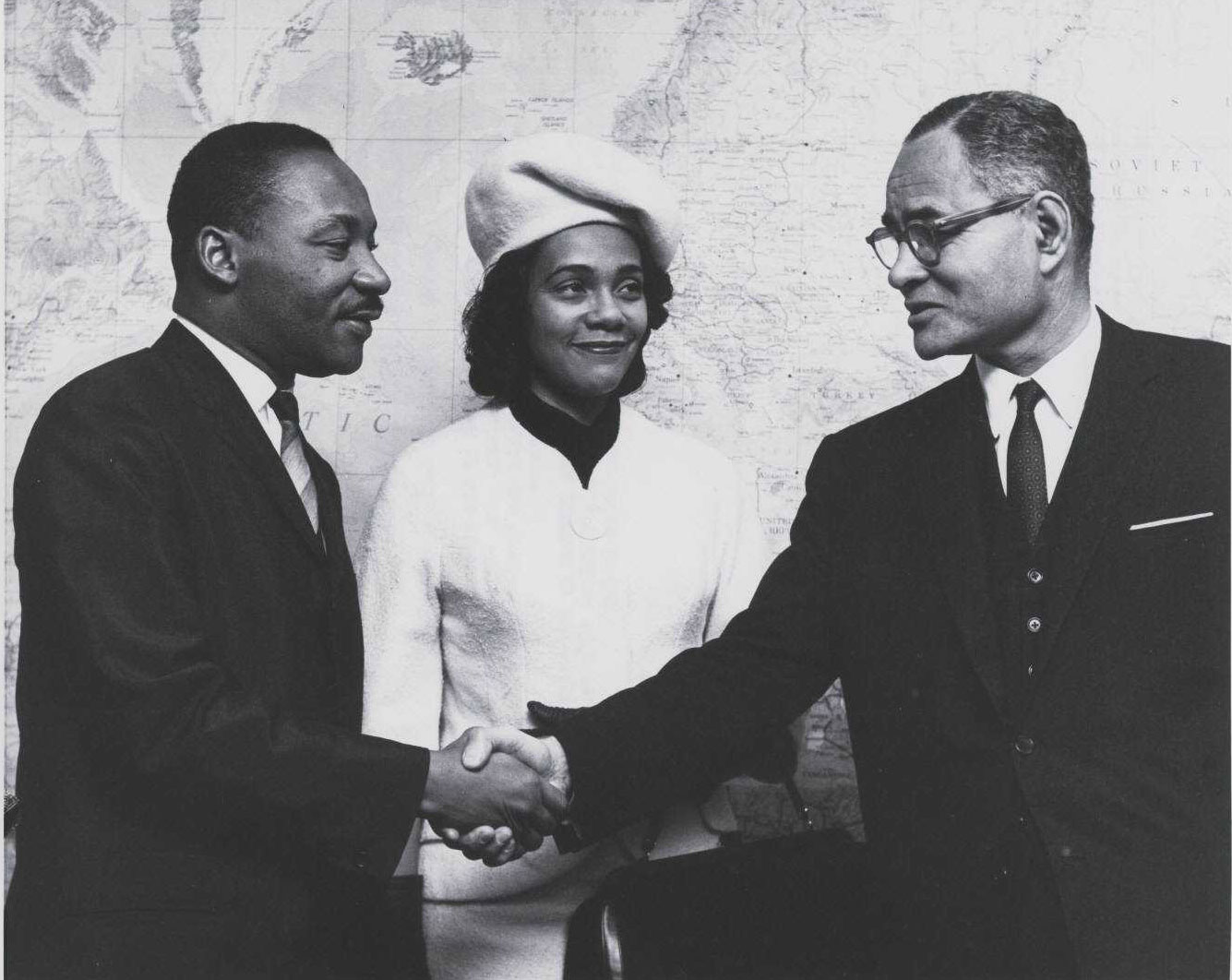 A black-and-white photograph of Martin Luther King Jr. and Ralph Bunche shaking hands, with Coretta Scott King standing between them, in front of a large world map.