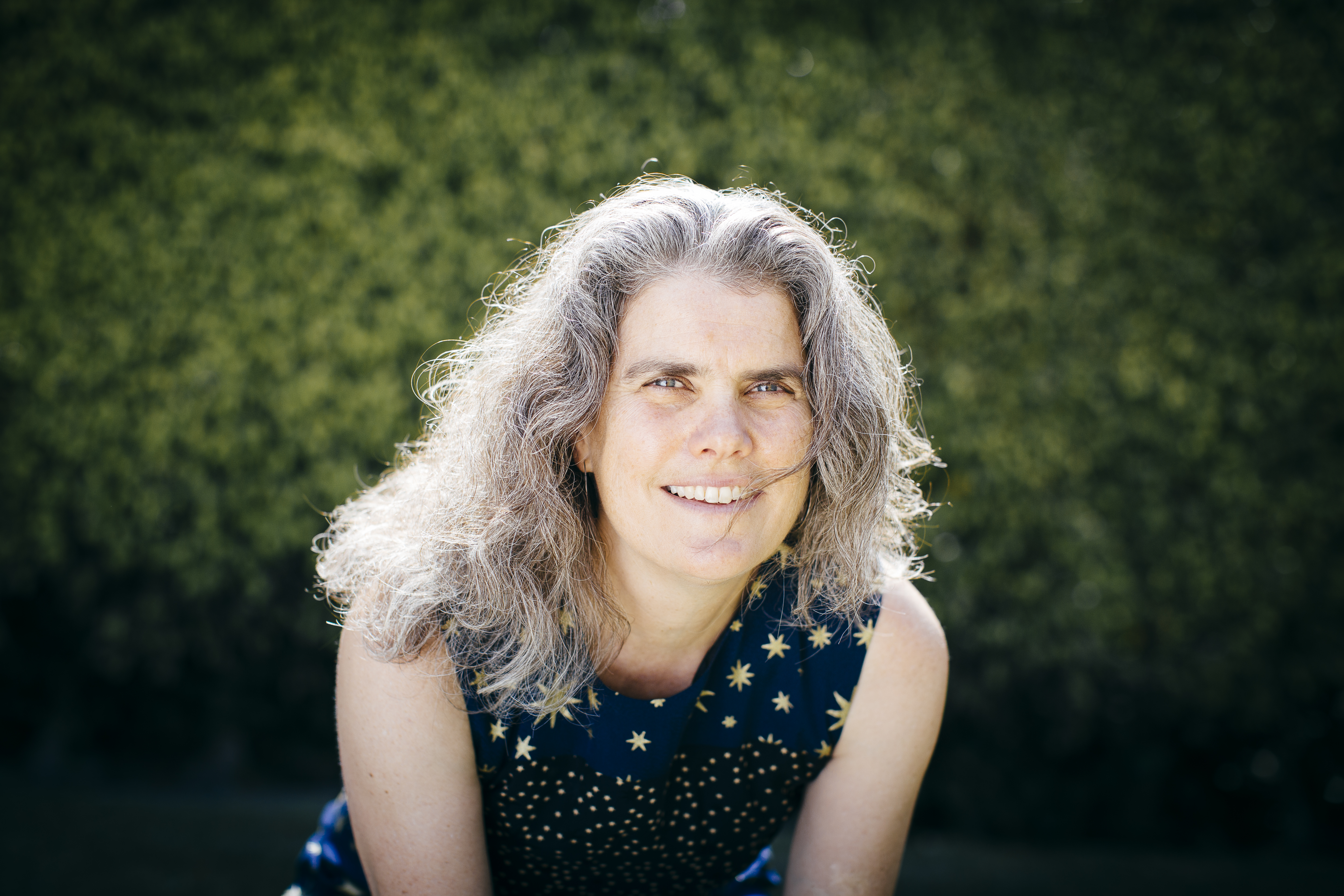 Andrea Ghez (head and shoulders) in a blue top with gold stars leans forward and smiles, with foliage in the background